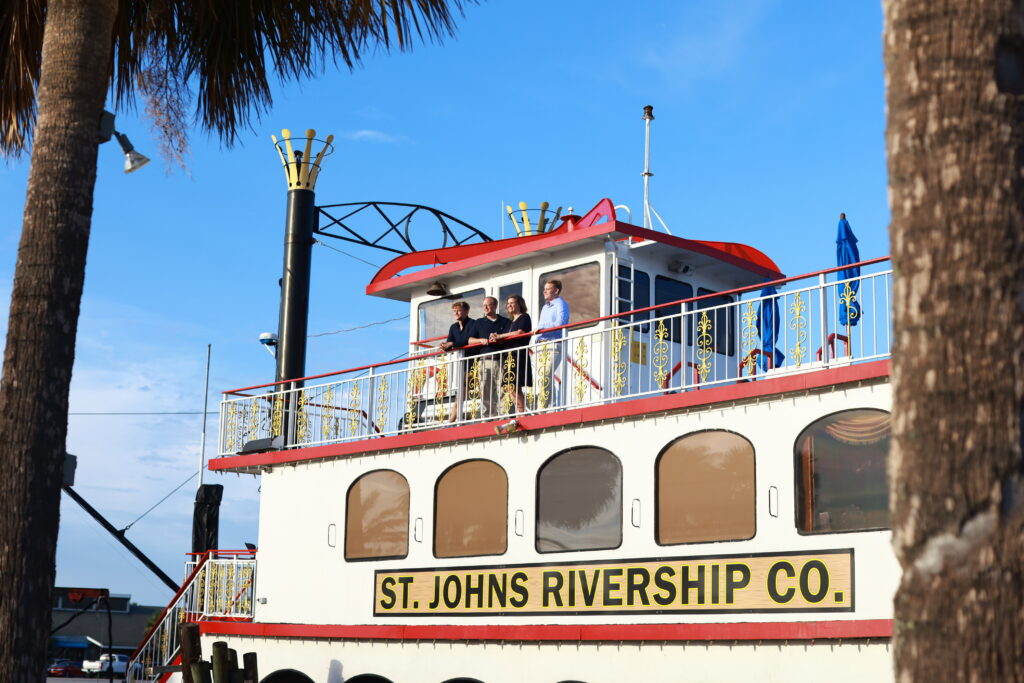 A family of four are standing on top of a steam boat. The boat has a banner that reads "St. Johns Rivership Co."
