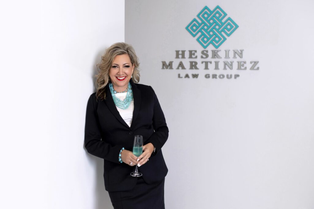 An ashy blonde woman is standing with a glass of teal champagne in front of a white wall that says "Heskin Martinez." She's wearing a teal necklace and a black suit.