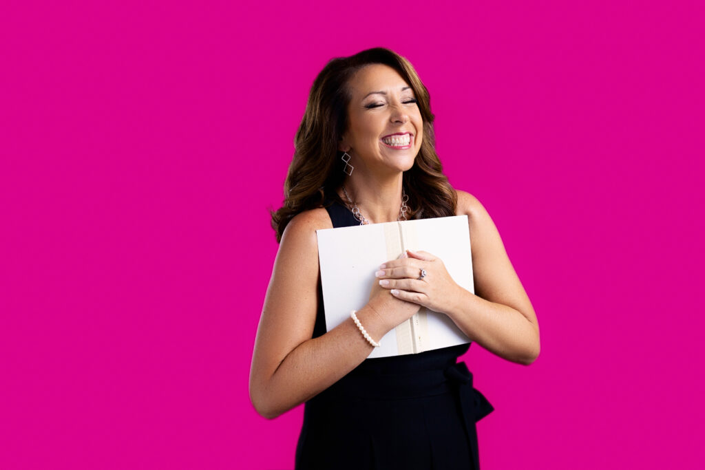 A brunette woman is holding a white book to her chest and grinning with her eyes closed. 