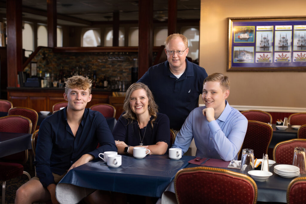 A family of four sits at a dinner table. The father is standing behind the mother and sons who are seated and sipping coffee.