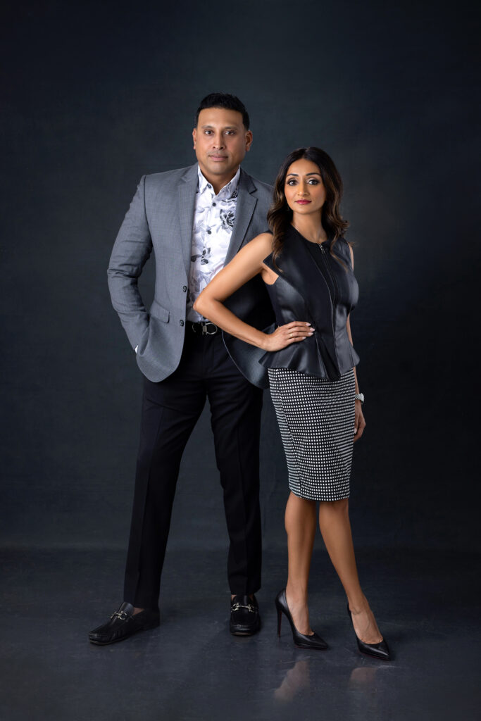 An Indian couple dressed in business formal outfits pose together in front of a black backdrop. The man is wearing a gray suit jacket and a white shirt and the woman is wearing a black blouse and checkered black and white pencil skirt.