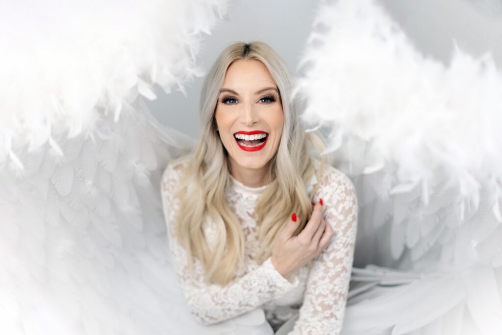 A blonde woman is grinning in a white, lace shirt while wearing white angel wings.