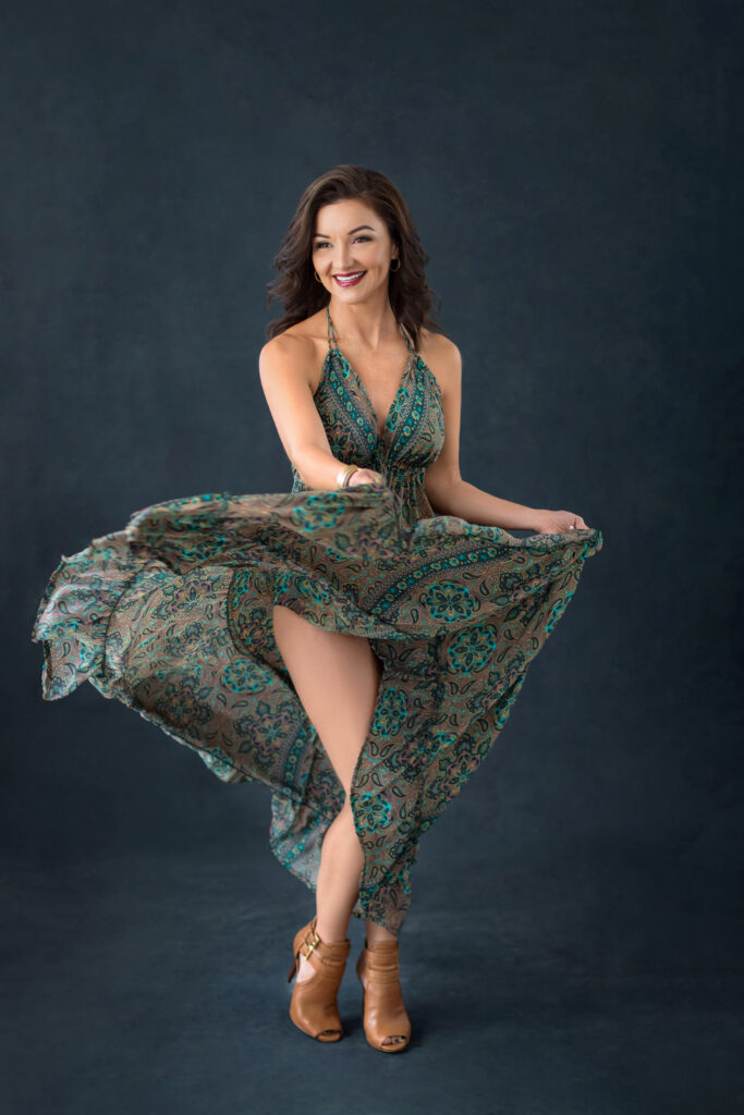 A brunette woman is grinning while she swishes her teal and light brown dress around in the air.