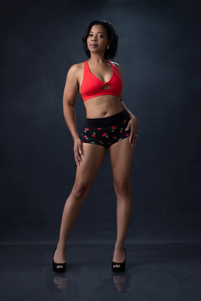 An African-American woman stands in front of a black backdrop wearing a red and black bathing suit. 